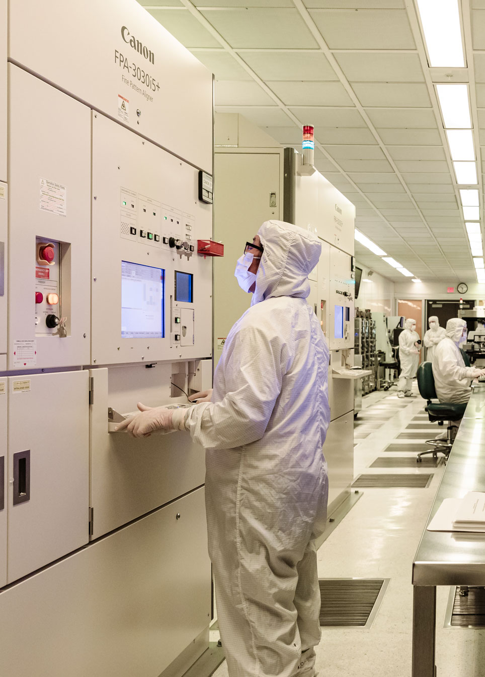 A technician in a cleanroom wearing a white lab coat and face mask operates advanced microelectronics manufacturing equipment. The background shows additional staff engaged in research, highlighting the precision and innovation in Northrop Grumman's microelectronics solutions.