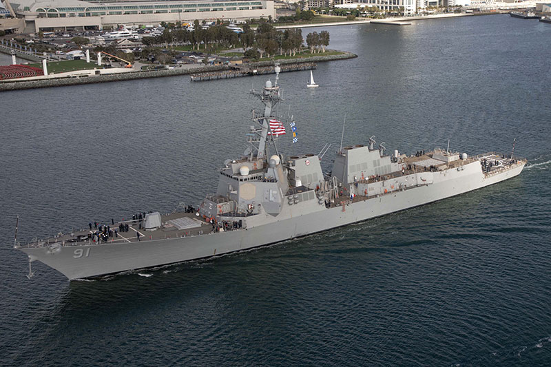 U.S. Navy destroyer USS Primetime 91 sailing near coastal city harbor with American flag visible.