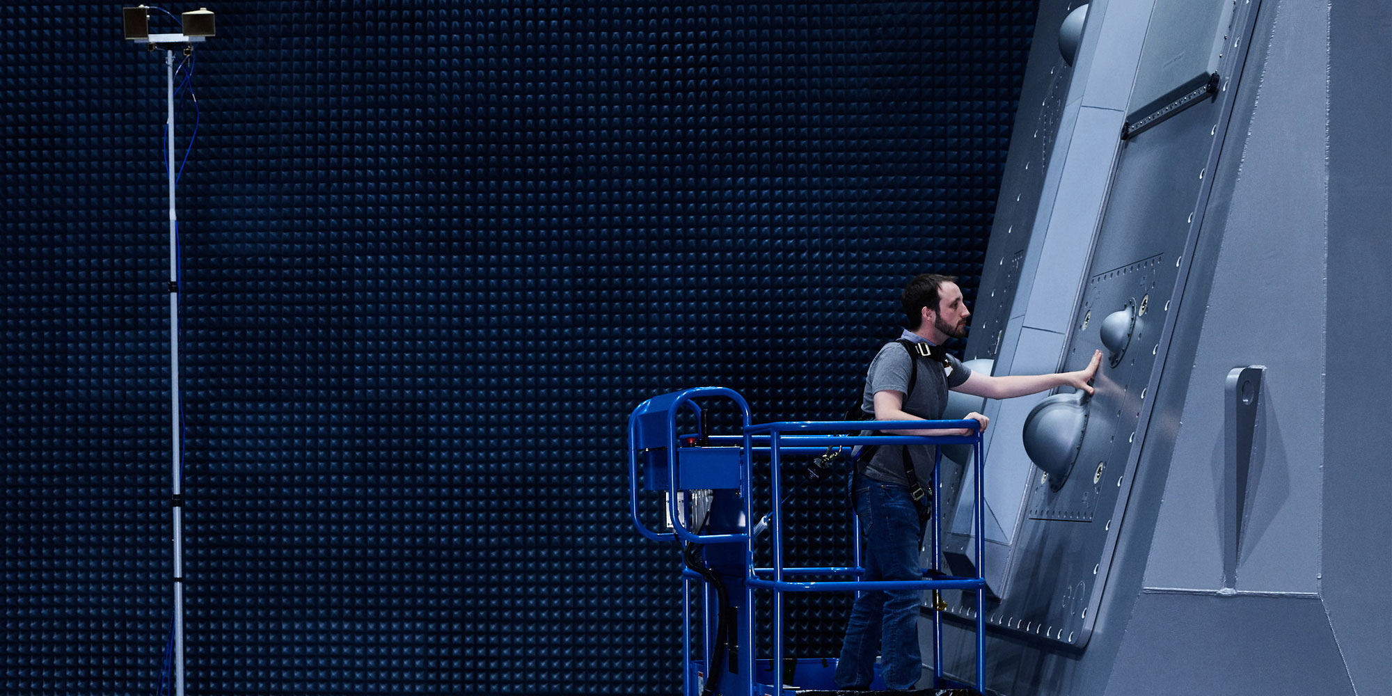 Surface Electronic Warfare Improvement Program (SEWIP) Technician inspecting naval radar system in anechoic chamber using blue safety lift platform indoors.