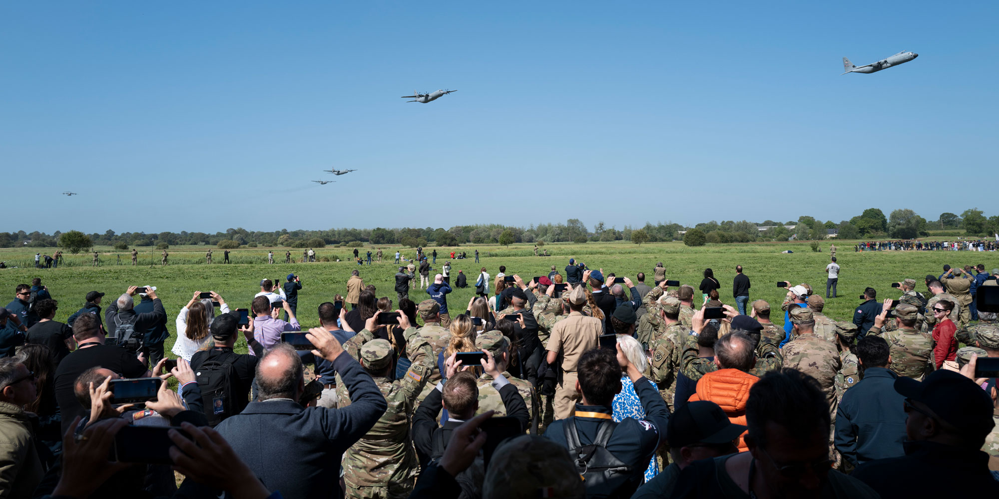 A crowd of people on a green grass airfield photograph five planes flying overhead