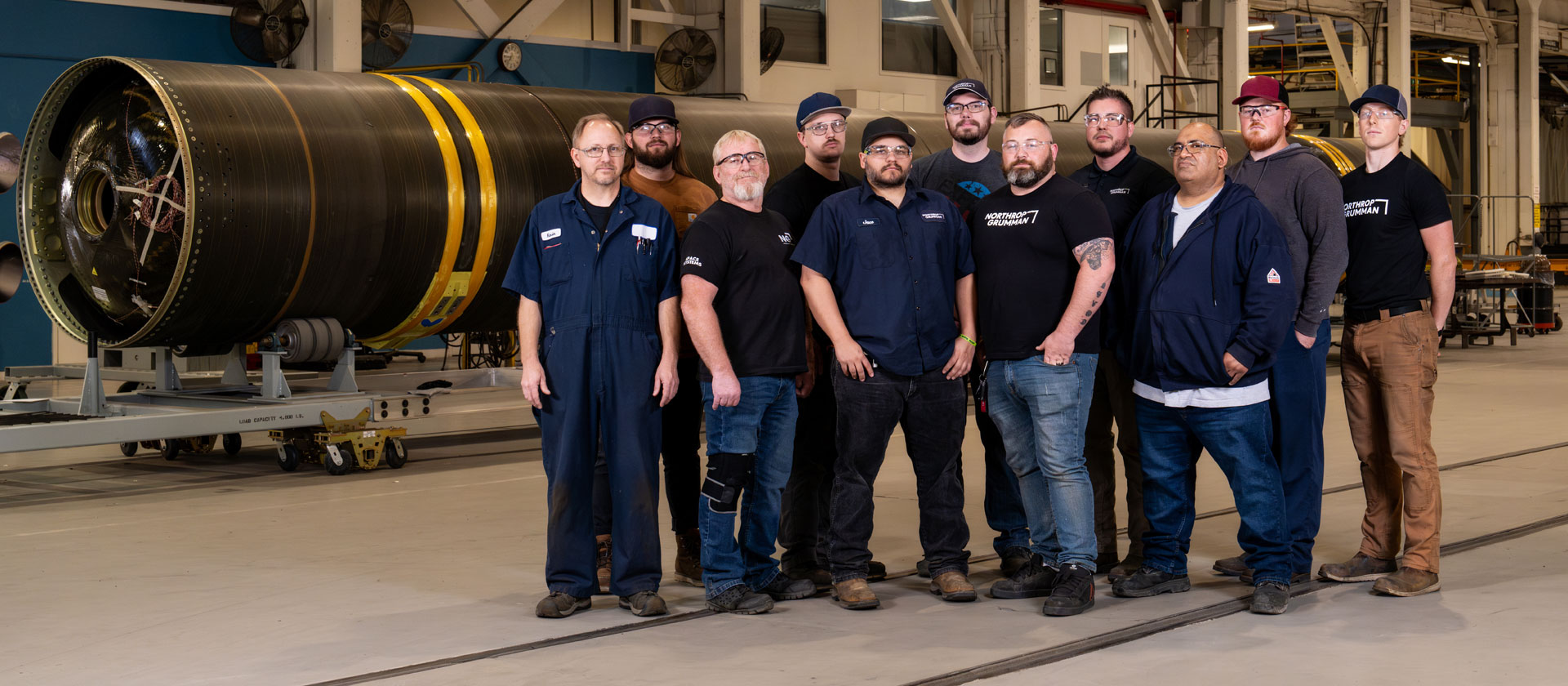 Group photo of Northrop Grumman hydrotesting team standing in a manufacturing facility, with a large solid rocket motor case in the background.