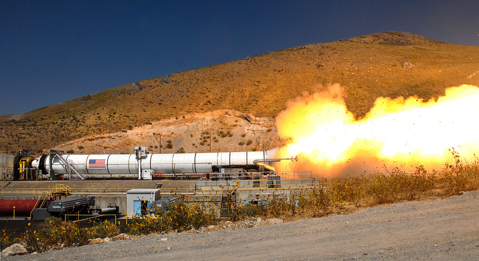 DM 3 Test Fire for Aretmis Mission – Northrop Grumman Horizontal rocket moter in desert environment during test fire, with smoke and flames coming out of the back of rocket.