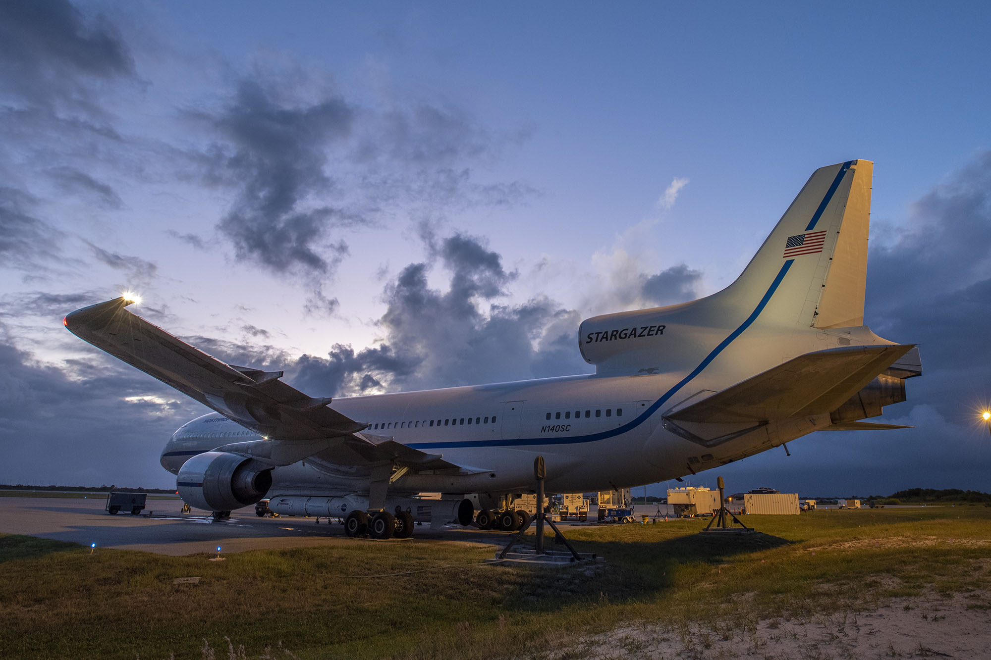 Pegasus_L1011_Skidstrip Resize Rear side view of airplane parked off the side of a runway in front of dark blue sky at sunset