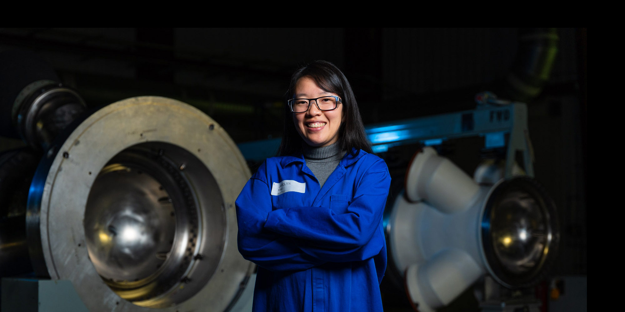 Jocelyn Chu – Materials and Process Design Engineer, Launch Abort System Abort Motor Asian female in blue lab coat and safety goggles stands in hallway of manufacturing plant